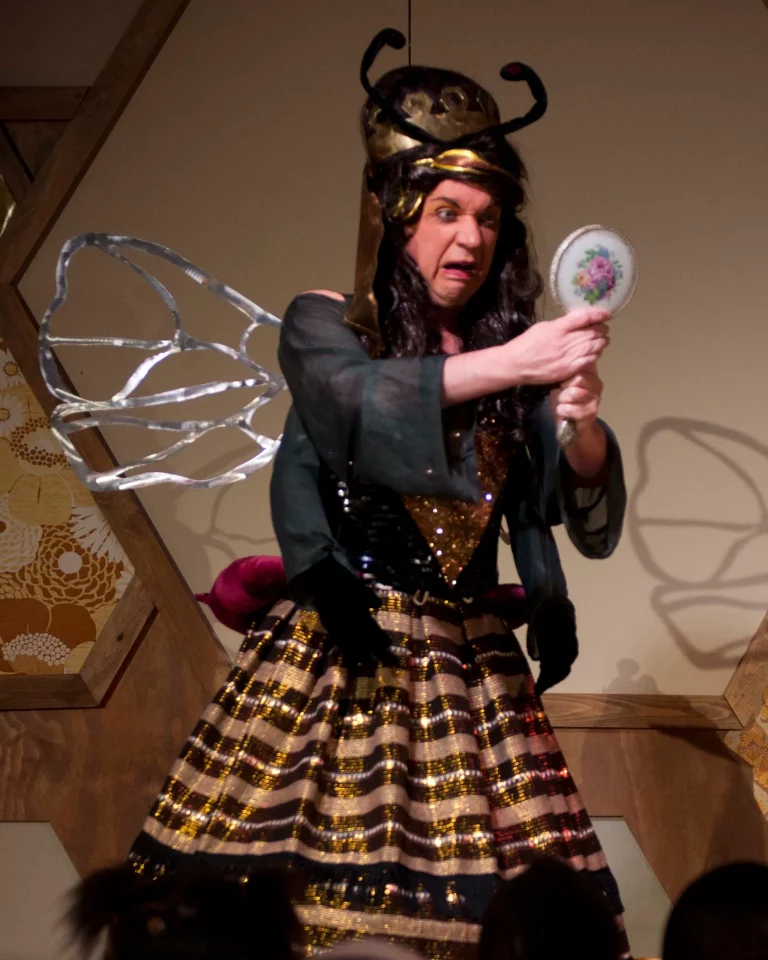Photo of the live show: a performer dressed as the queen bee, with antennae and silver wings, holding a hand mirror