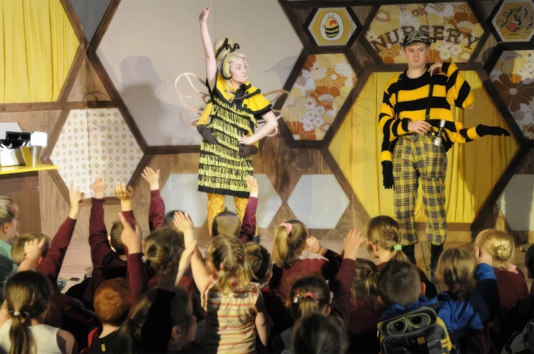 Photo of the live show: a performer in a bee costume on stage as children in the audience raise their hands