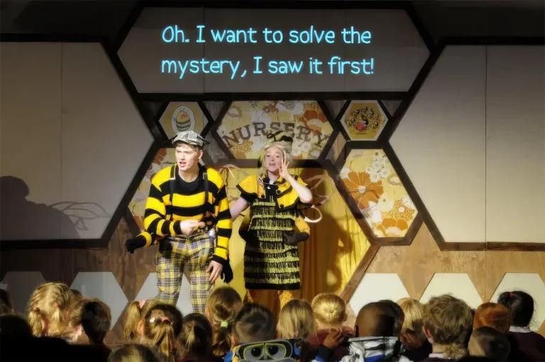 Photo of the live show: two actors in bee costumes on stage with projected honeycomb scenery and the caption “Oh, I want to solve the mystery, I saw it first!”, watched by children